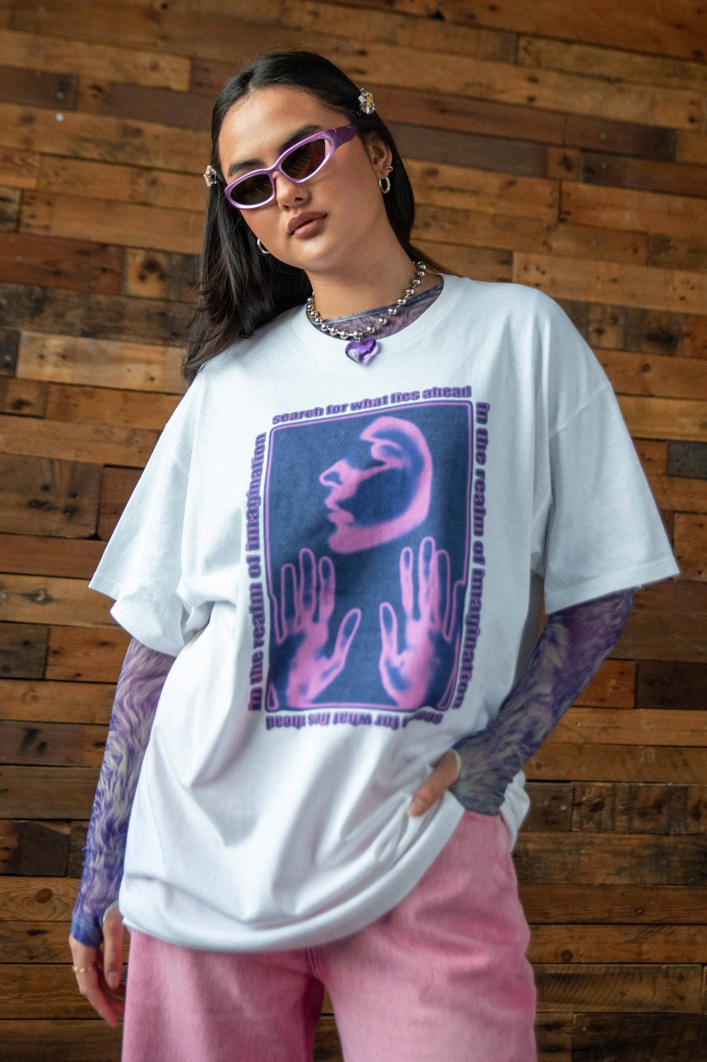 Unisex white cotton T-shirt featuring bold purple surreal graphic and 'Realm of Imagination' text, paired with pink trousers and purple mesh sleeves.