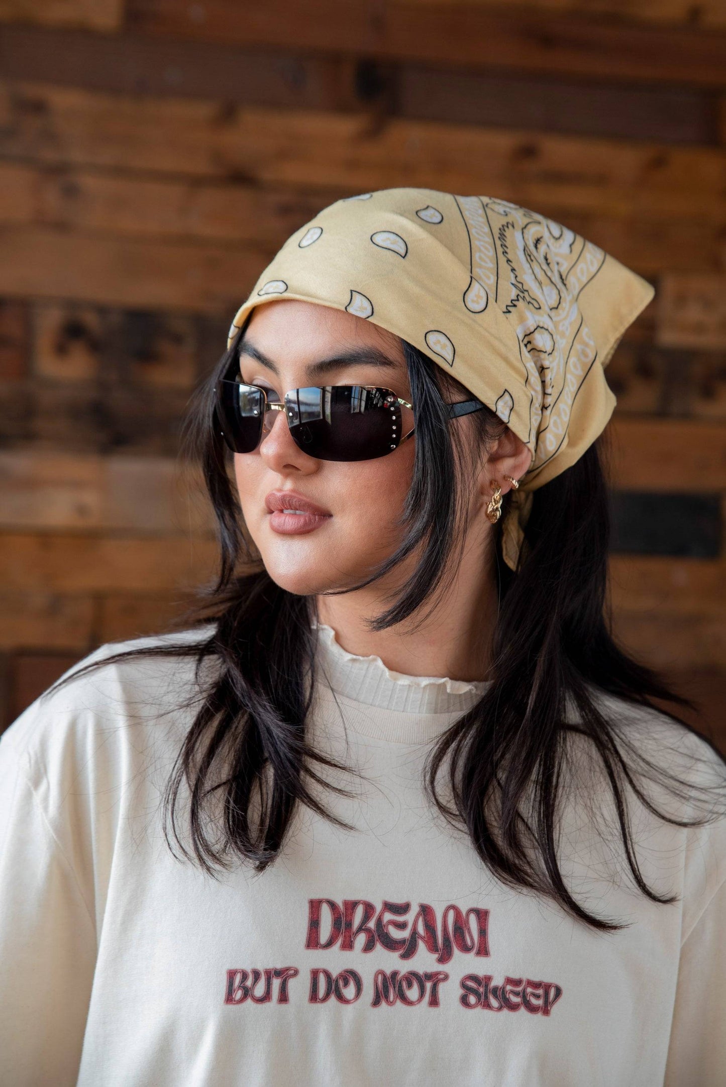 Model wearing natural cotton t-shirt with logo print, styled with sunglasses and a yellow bandana for a casual streetwear look.