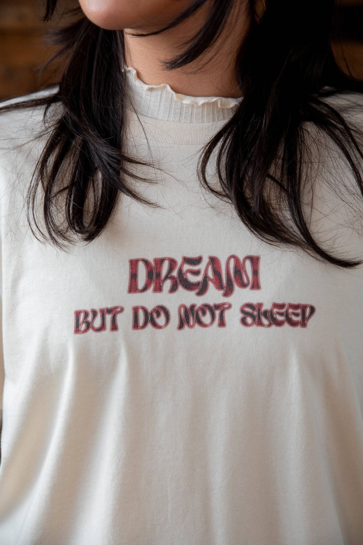 Close-up of natural cotton t-shirt showing bold red cursive logo print 'DREAM BUT DO NOT SLEEP' with soft fabric texture.
