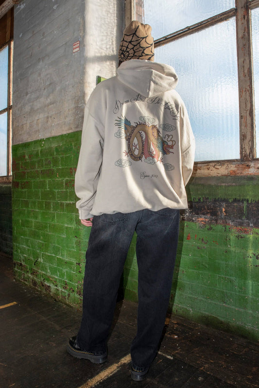 Back view of sand-colored hoodie featuring detailed Chinese dragon artwork and slogan, worn with black jeans in a streetwear setting.