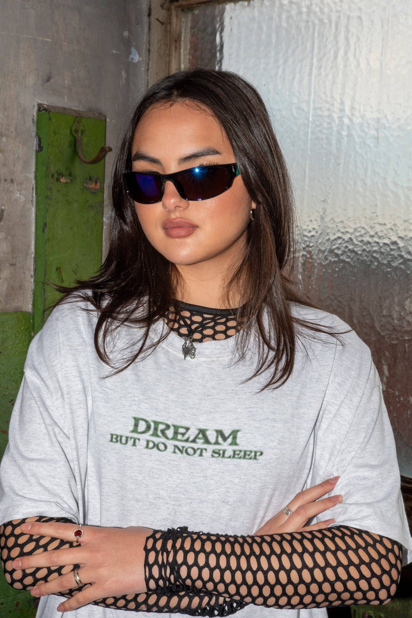 Close-up of ash grey short sleeve T-shirt featuring bold green futuristic logo print, layered over black fishnet top for edgy streetwear look.