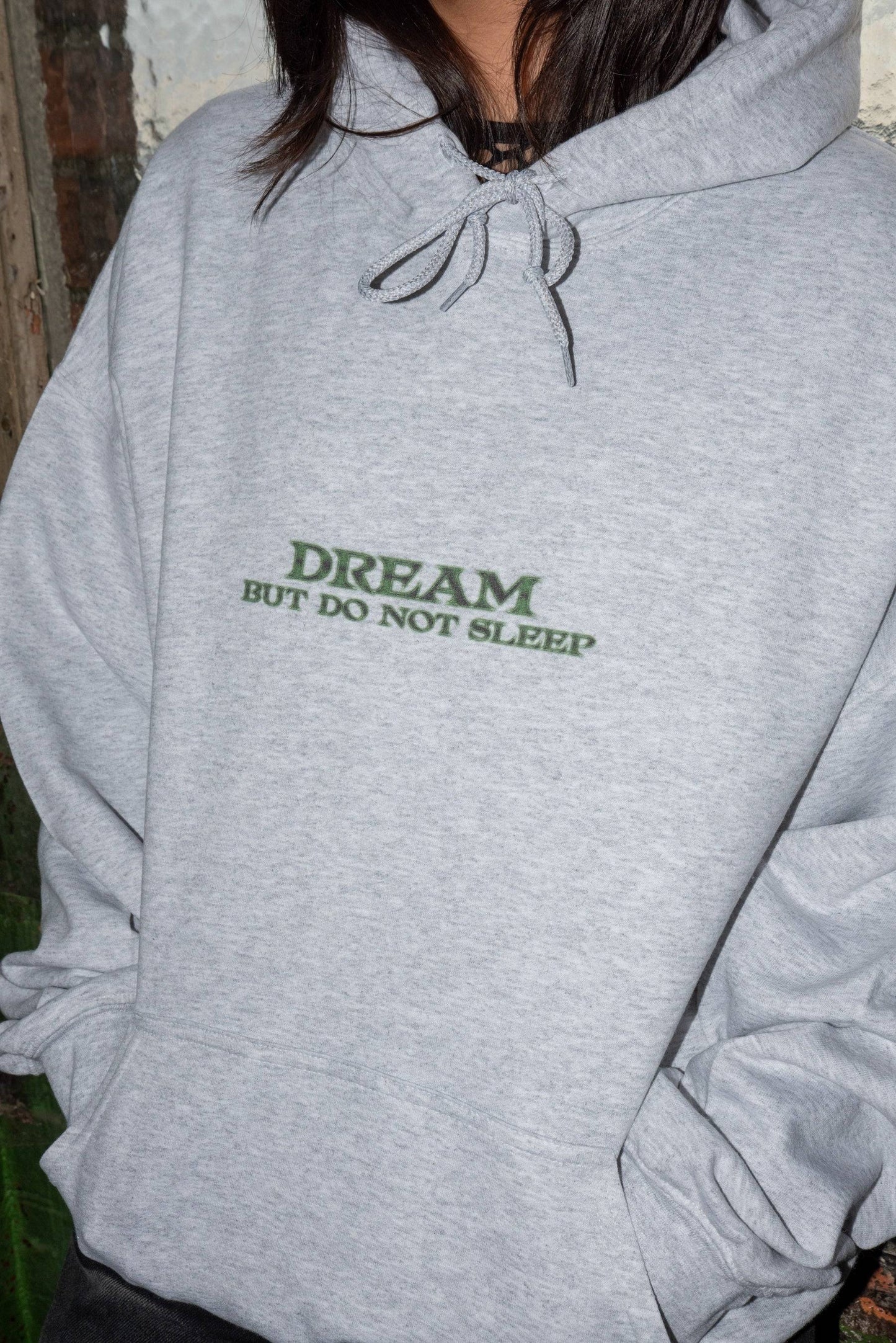 Detail view of DBDNS logo on ash grey hoodie reading 'DREAM BUT DO NOT SLEEP'. Relaxed fit cotton hoodie in streetwear style.