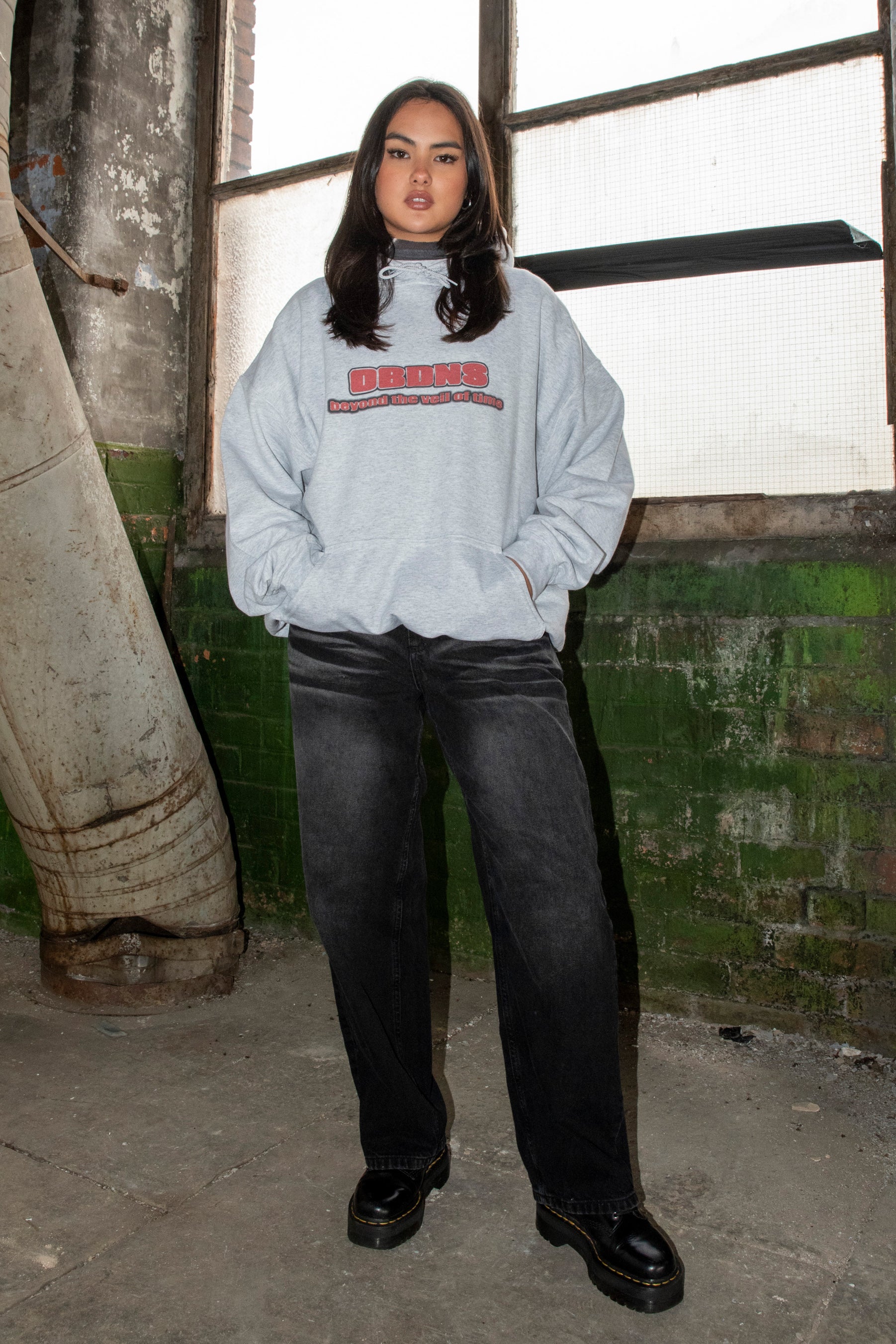 Full-body view of model wearing ash grey DBNDNS logo hoodie styled with black jeans, showcasing relaxed streetwear fit.