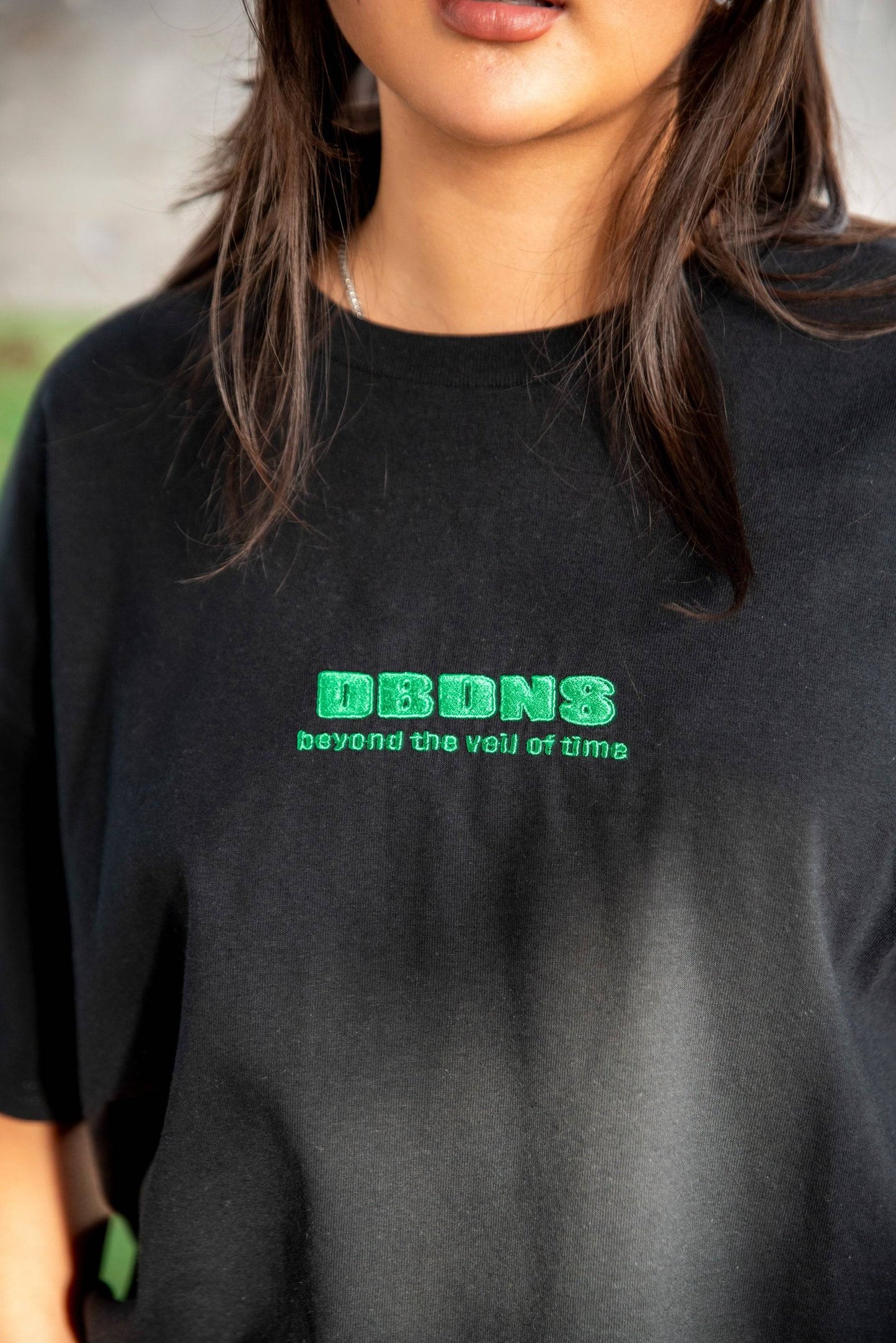 Close-up of black T-shirt showing green embroidered DBDNS logo with text 'beyond the veil of time'. 100% cotton short sleeved tee.