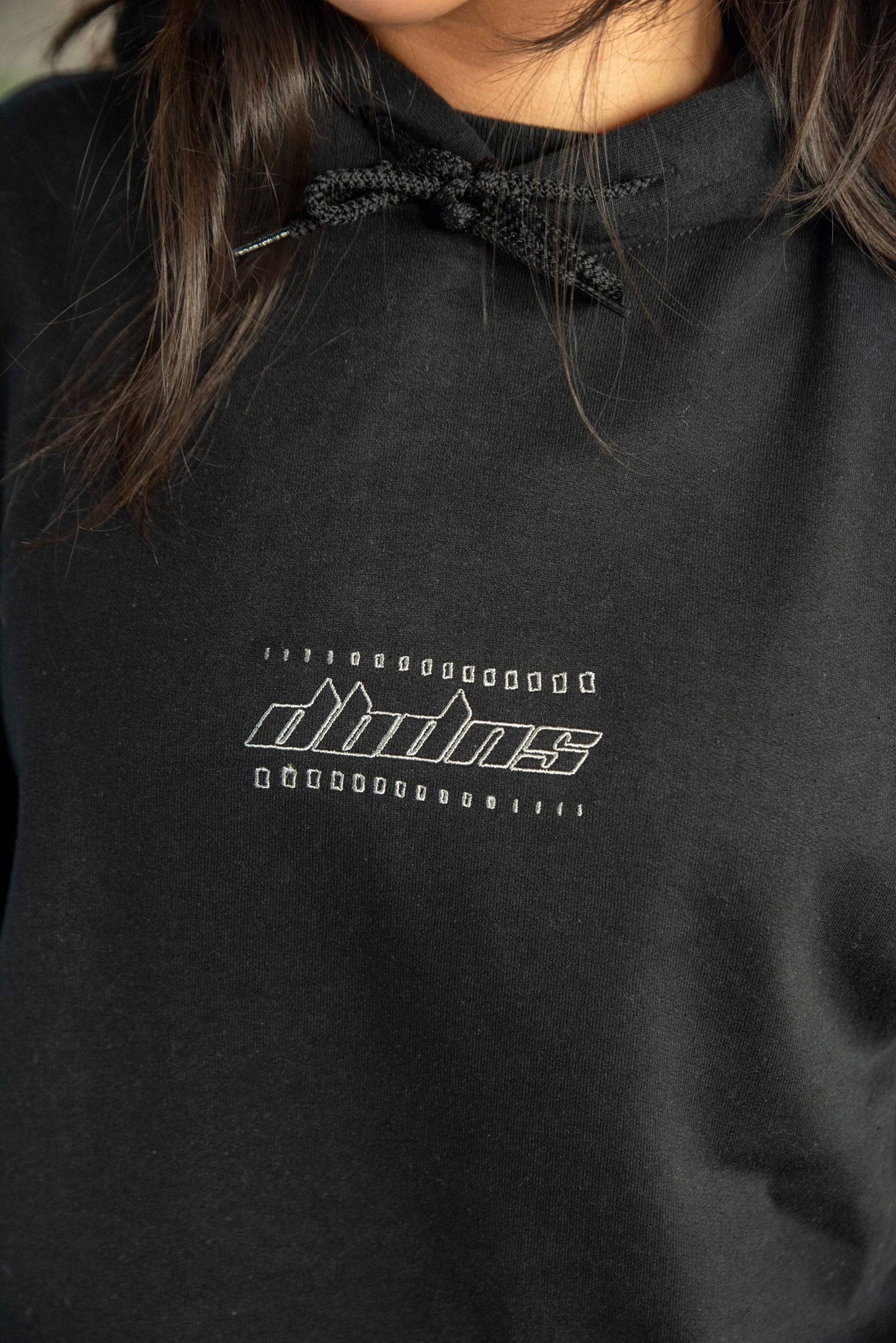 Close-up of black unisex hoodie featuring futuristic embroidered logo on chest. Heavyweight cotton fabric with drawstring hood detail.
