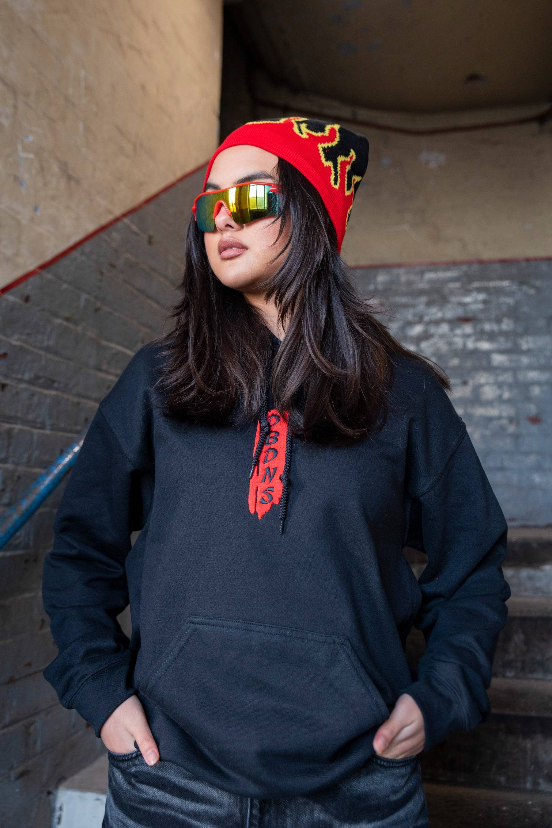 Unisex black hoodie with red dragon embroidery and DBDNS branding, styled with a red flame beanie and reflective sunglasses.