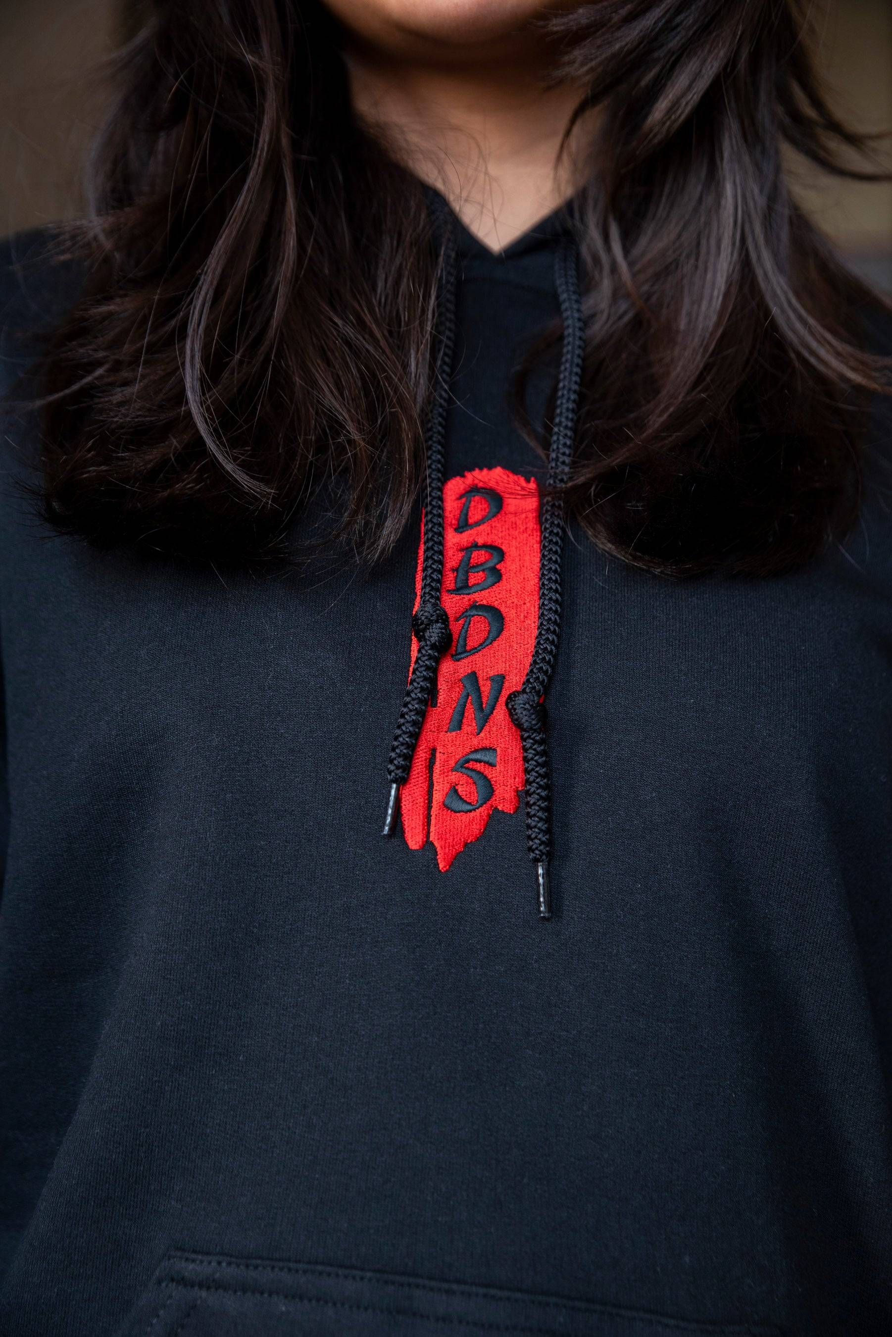 Close-up of DBDNS red embroidered logo on the drawstrings of the black hoodie, showcasing streetwear branding details.