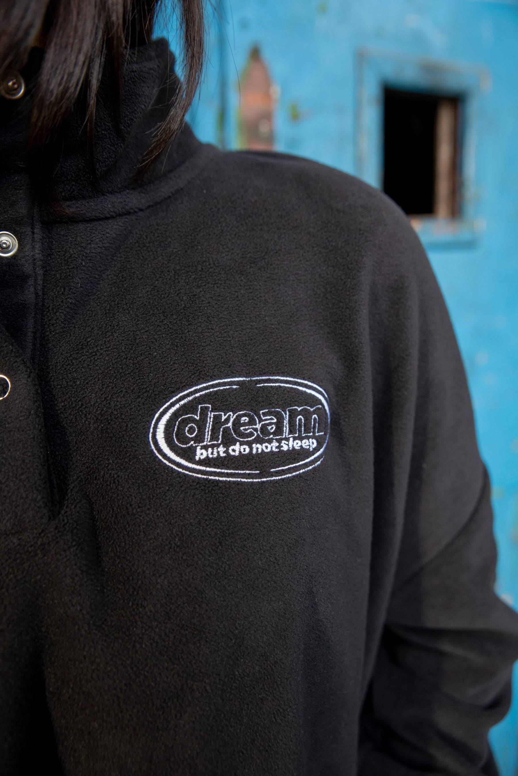 Detailed view of oval 'dream just do not sleep' logo embroidery on Black Cropped Fleece Sweatshirt. High-quality stitching on cozy fleece.