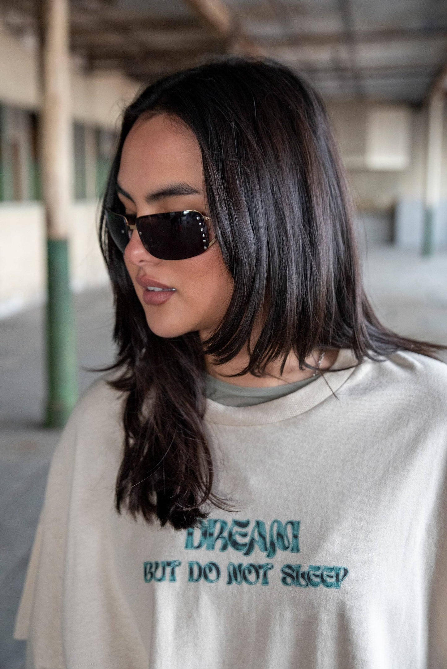 Model wearing Short Sleeved T-Shirt in Sand - Unisex Cotton Tee with bold cursive logo print, styled with sunglasses for a casual streetwear look.