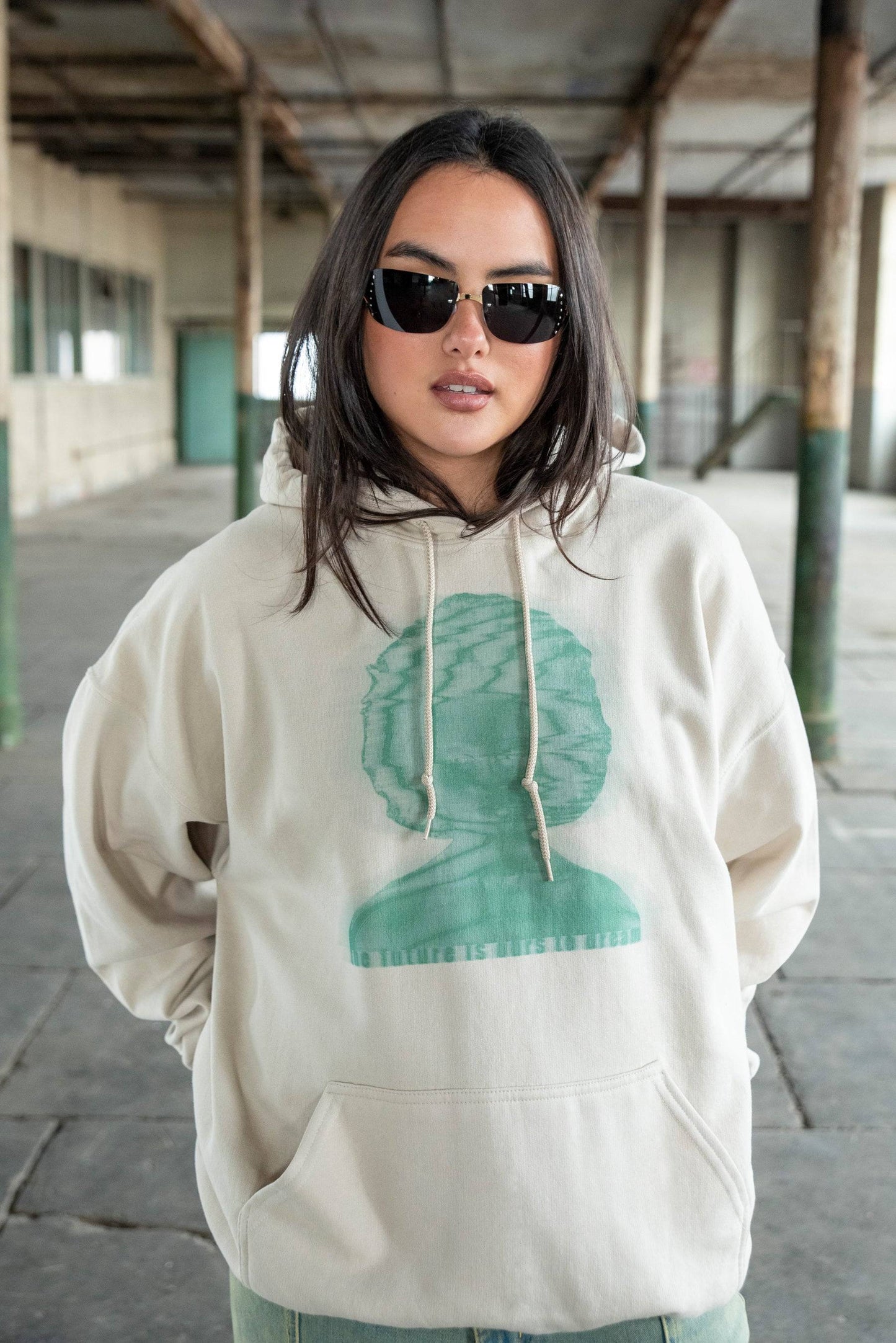 Boxy fit sand hoodie with green graphic and slogan, styled with sunglasses for a modern Y2K streetwear vibe in industrial backdrop.