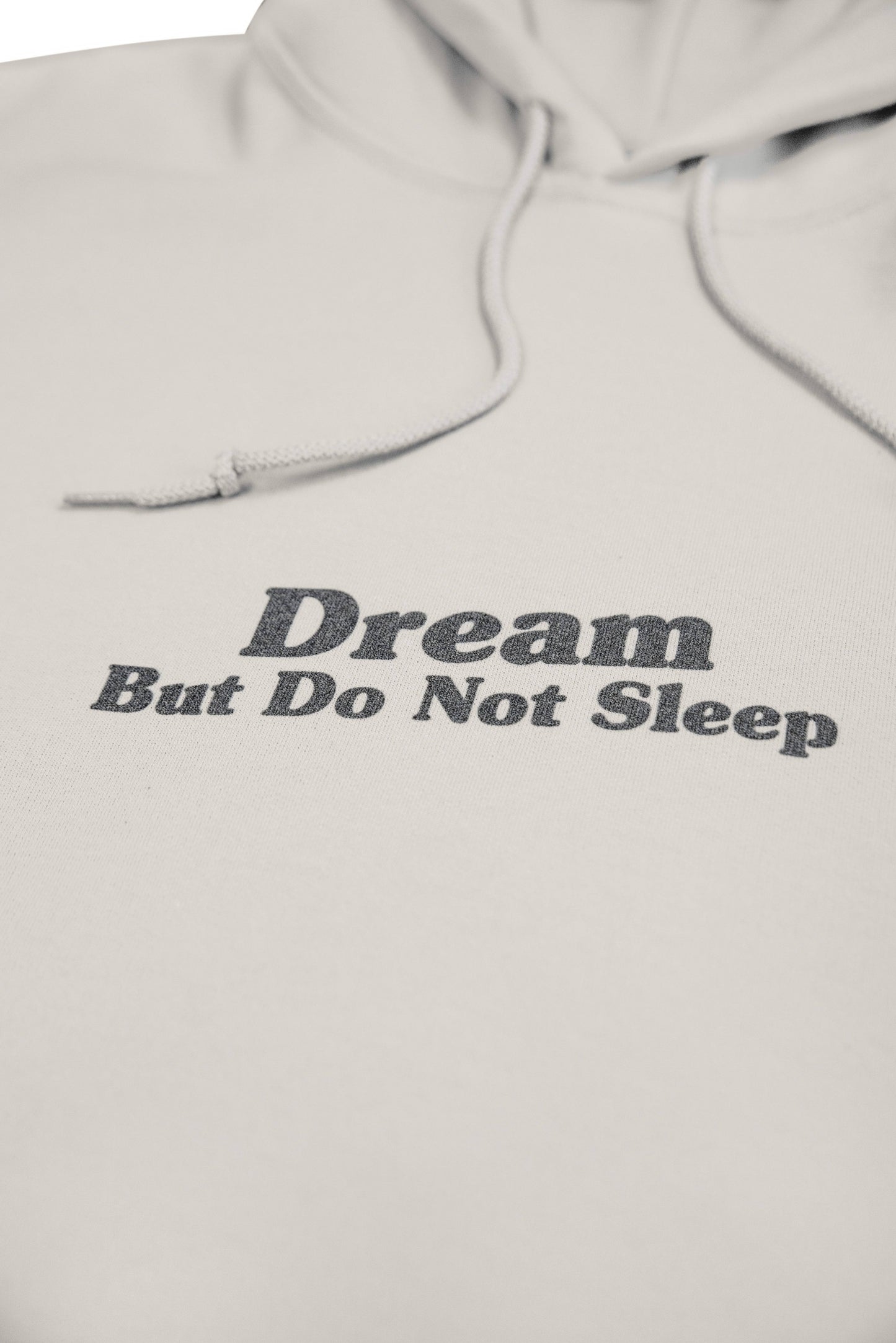 Macro shot of 'Dream But Do Not Sleep' text on sand hoodie. Soft heavyweight cotton fabric with subtle texture and clean stitching.