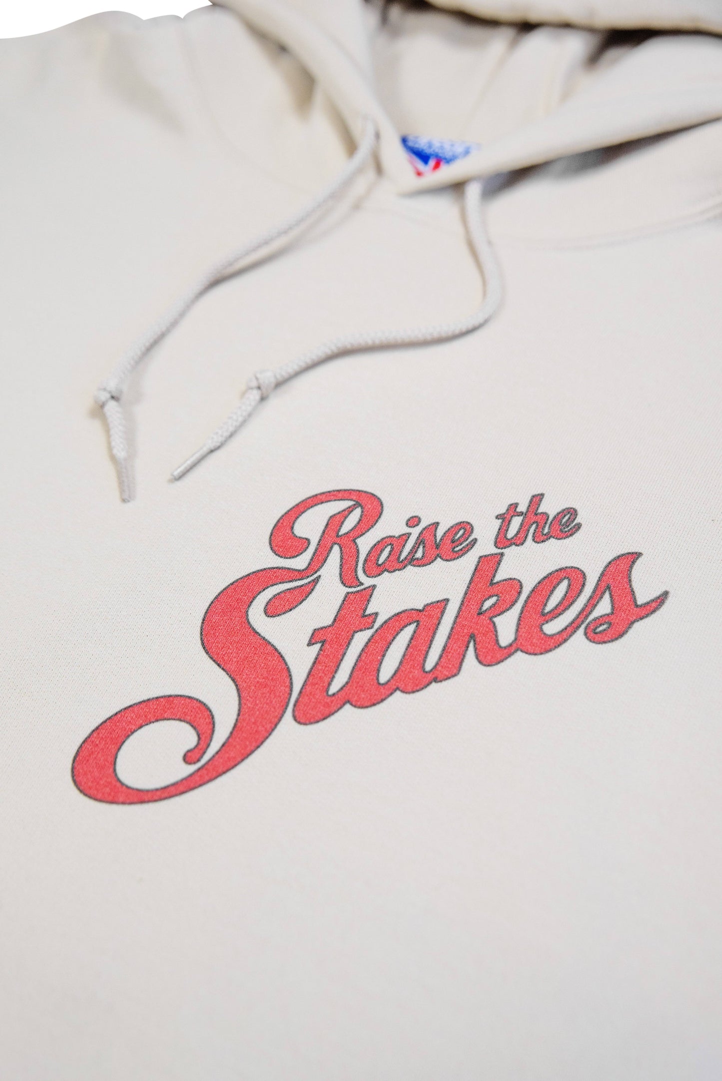 Angled close-up of chest logo on unisex sand hoodie with vintage-style 'Raise the Stakes' lettering in red.