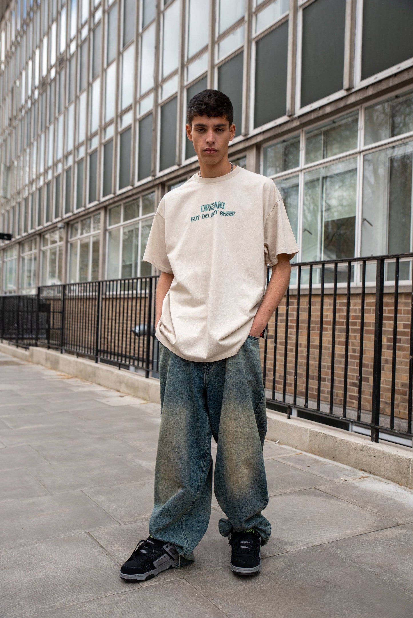 Unisex relaxed fit short-sleeved sand T-shirt with 'Dream But Do Not Sleep' logo, styled with baggy jeans in urban setting.