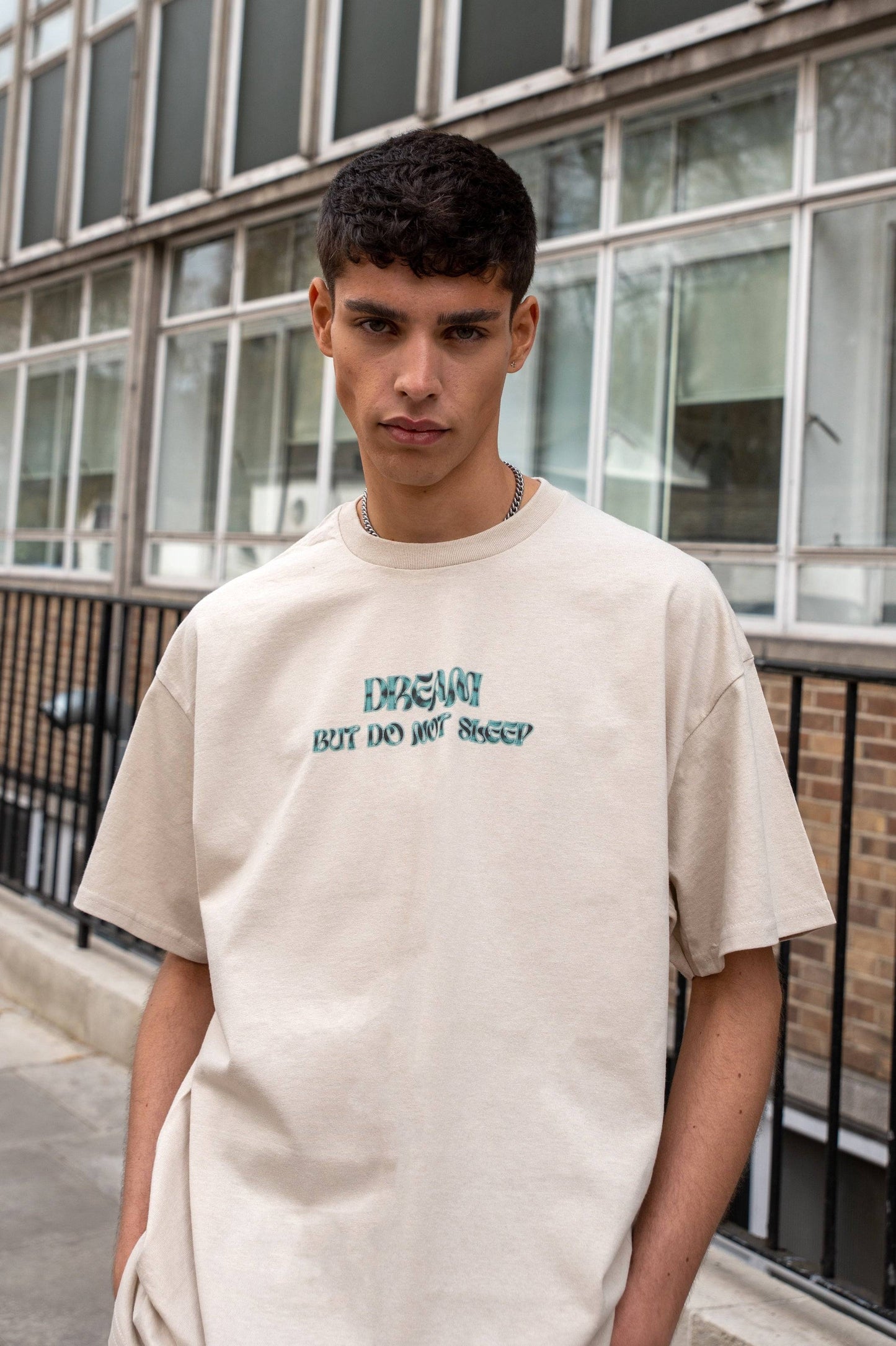 Close-up of sand short-sleeved T-shirt featuring green cursive 'Dream But Do Not Sleep' logo, part of unisex relaxed fit collection.