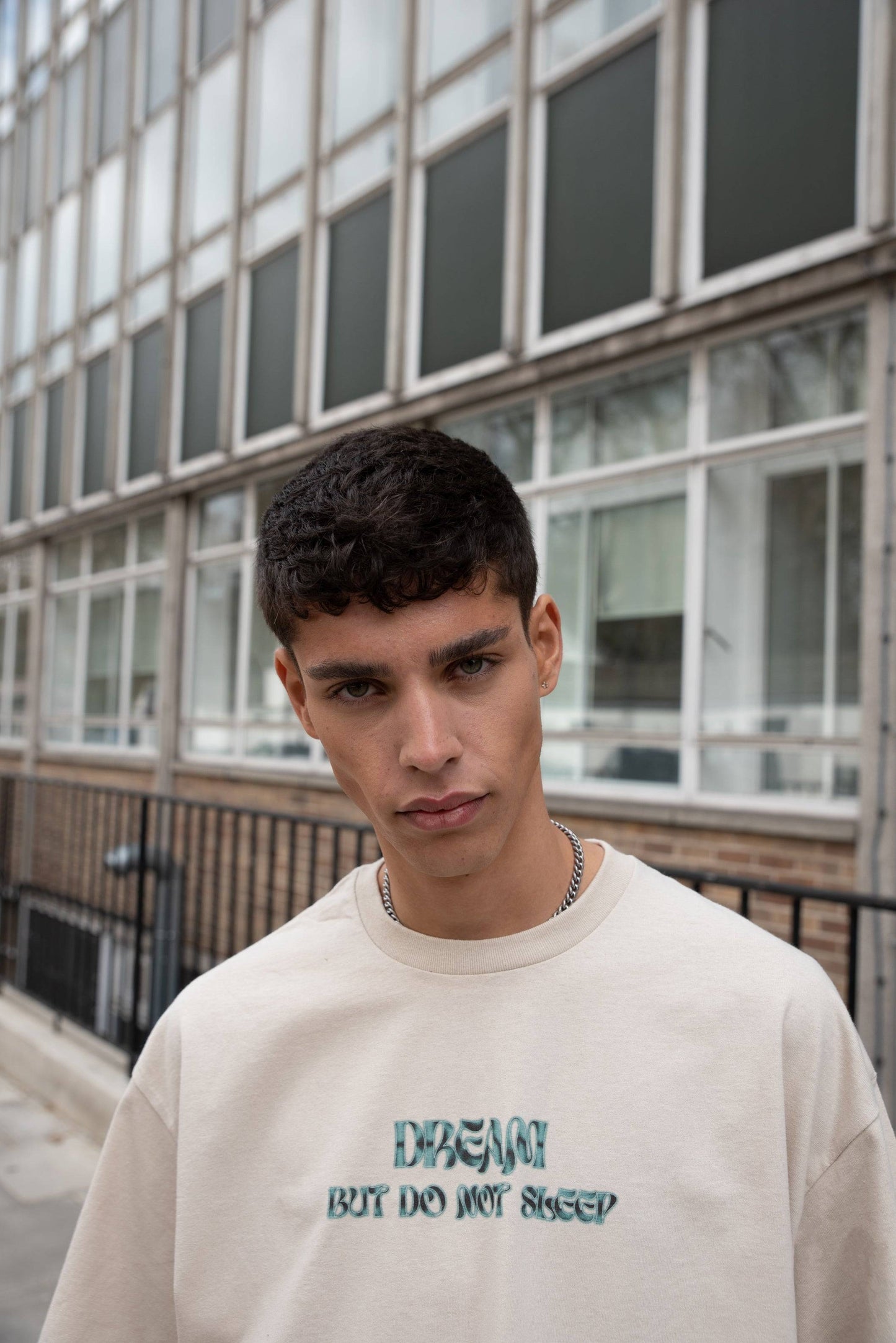 Model wearing short-sleeved sand T-shirt with bold green logo text, showcasing unisex relaxed fit streetwear style.