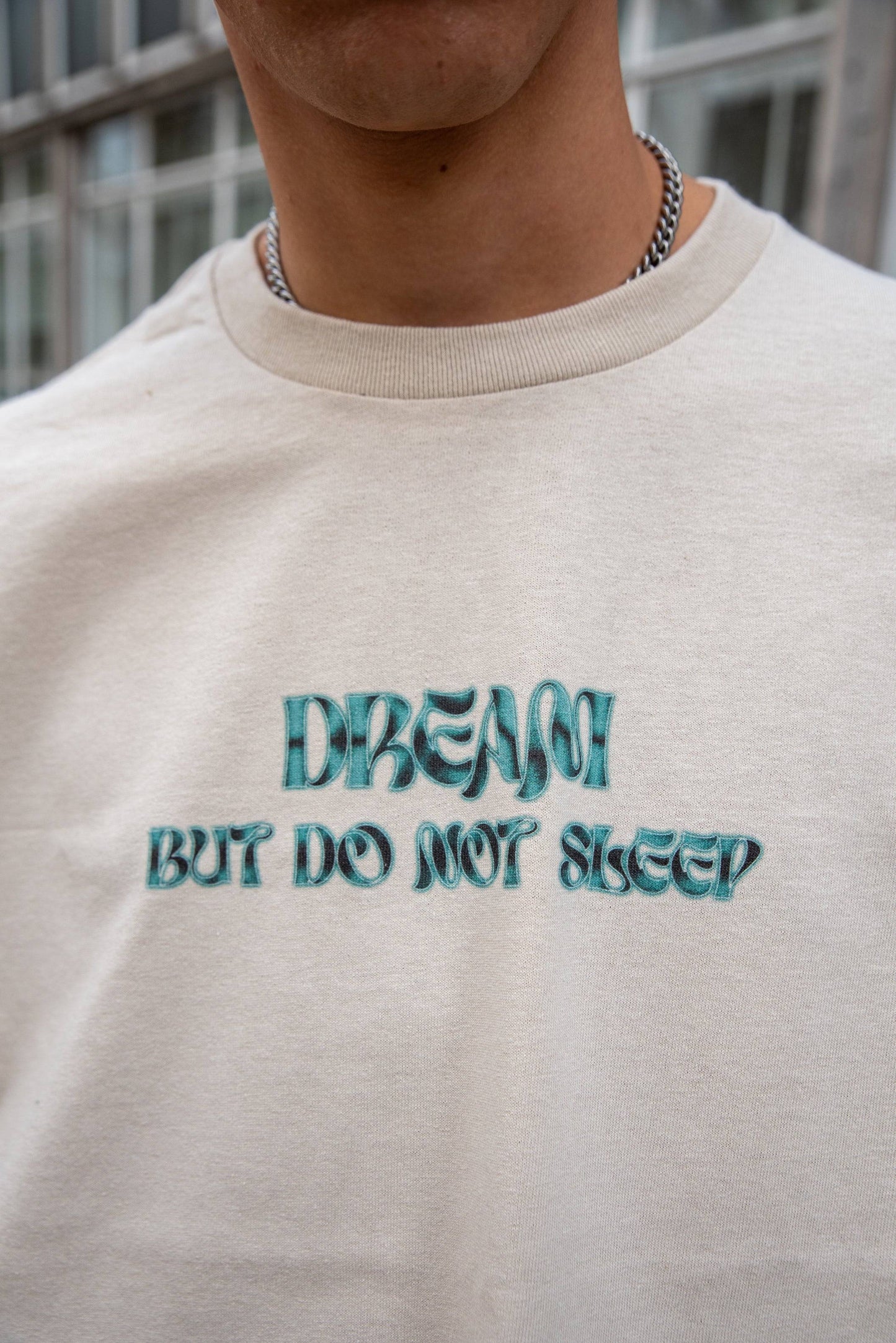 Detailed view of 'Dream But Do Not Sleep' logo on sand short-sleeved T-shirt, highlighting graphic print and fabric texture.