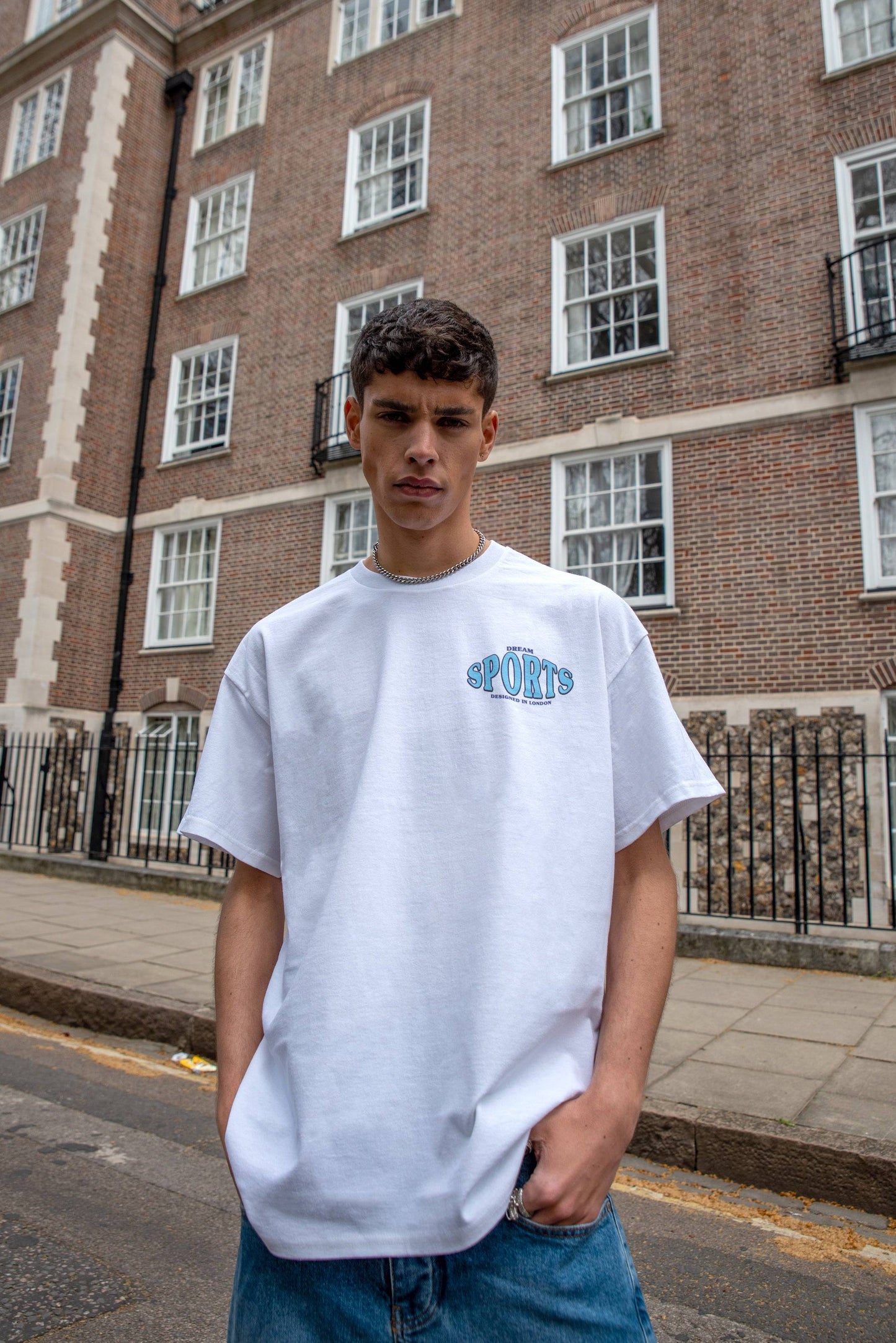 Front view of white short sleeved t-shirt featuring blue 'Dream Sports' chest print, styled with relaxed fit for streetwear fashion.