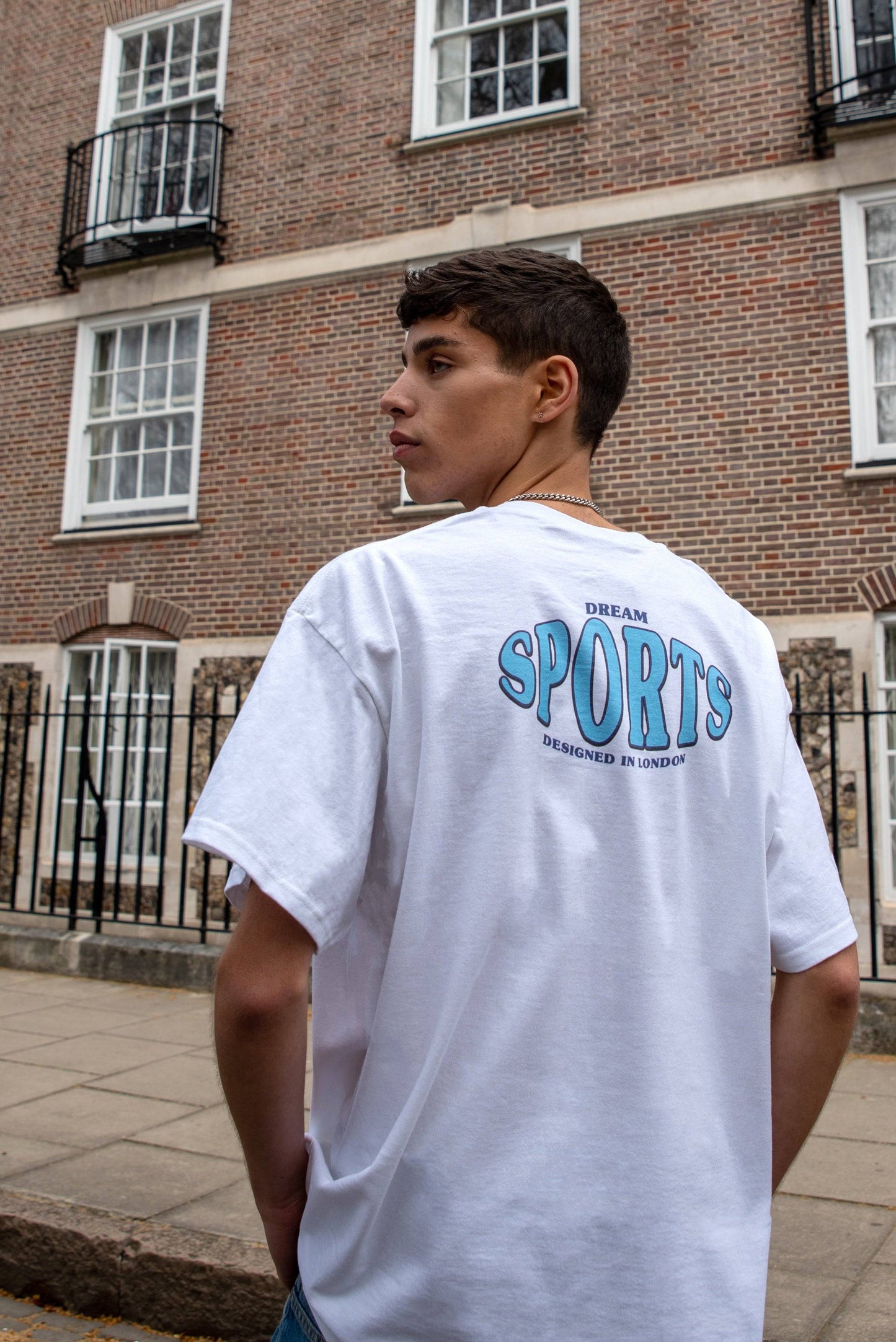 White sport print t-shirt with 'Dream Sports Designed in London' text on back, styled in urban streetwear setting. Regular fit short sleeve tee.