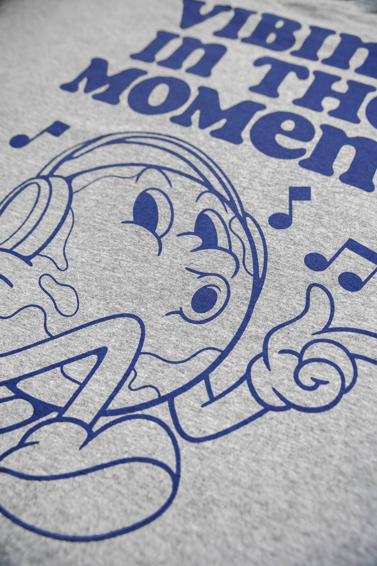 Close-up of cartoon character and 'Vibing in the Moment' text on grey short sleeve t-shirt. Durable blue ink on soft cotton fabric.