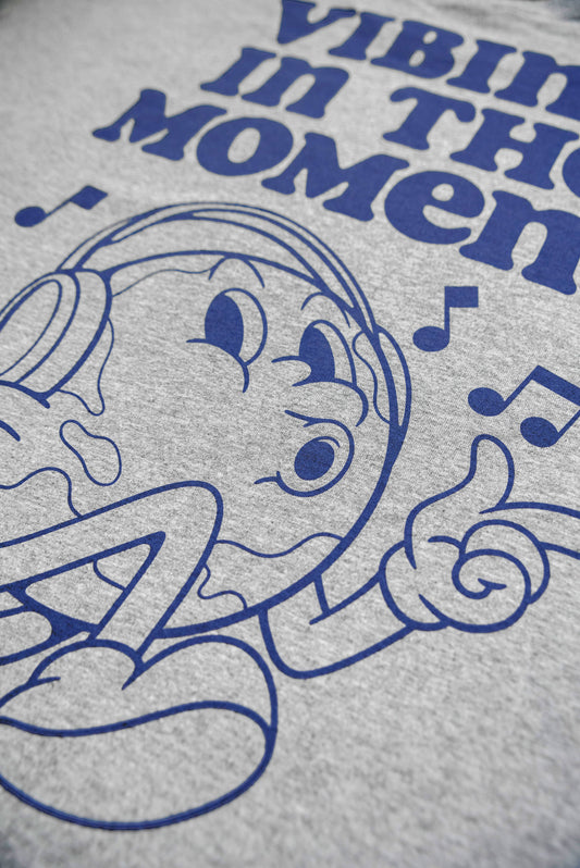 Close-up of cartoon character and 'Vibing in the Moment' text on grey short sleeve t-shirt. Durable blue ink on soft cotton fabric.