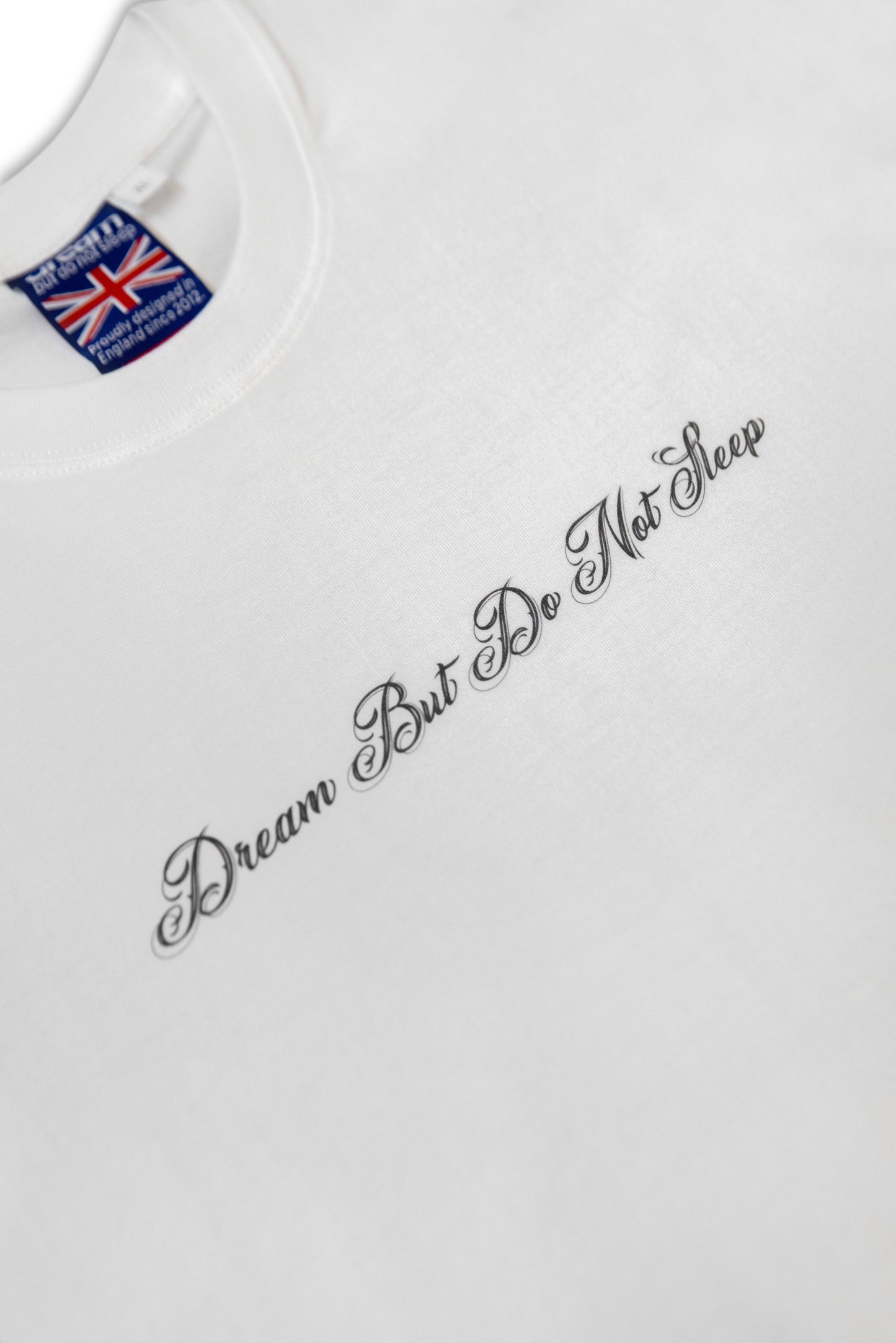 Close-up of the White Phoenix T-Shirt front text reading 'Dream But Do Not Sleep' in elegant script. Soft cotton fabric with precise lettering.