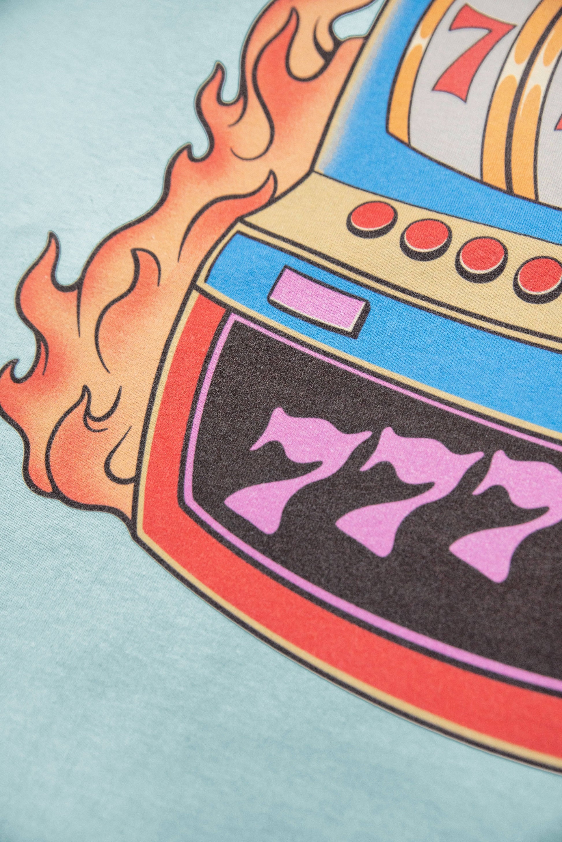 Zoomed-in bottom section of Lucky No 7 slot machine print on blue t-shirt, showcasing bold 777 and flame accents.