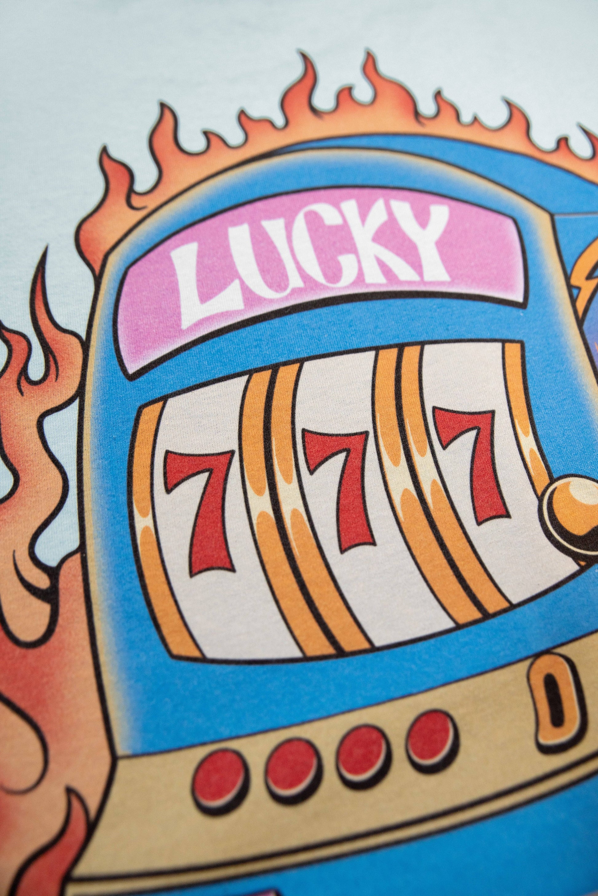 Close-up of Lucky No 7 slot machine graphic on blue short sleeved t-shirt, showing triple 7s and bold flame design.